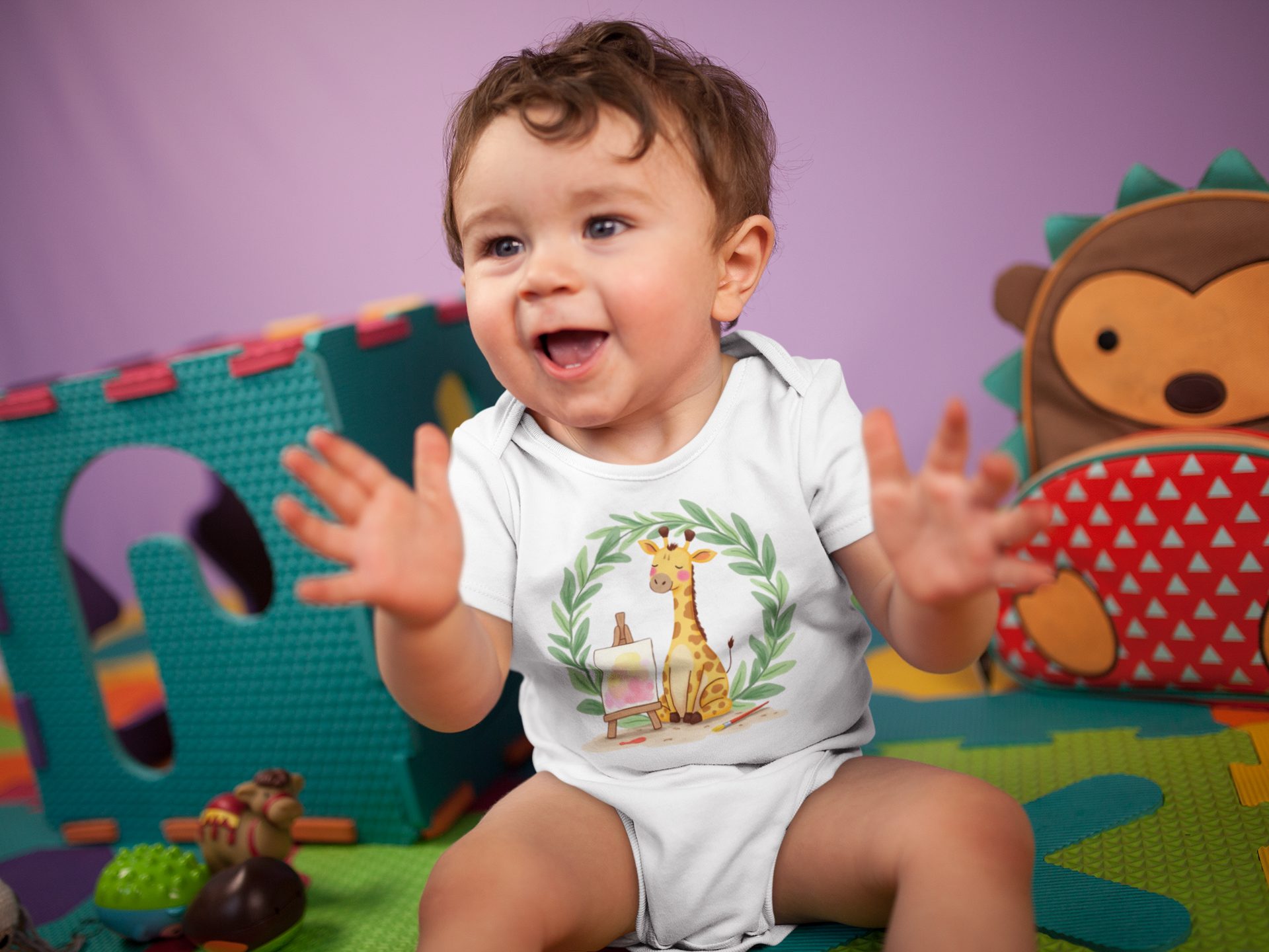 Baby sitting on a colorful play mat with toys, wearing a Little Chaos Crew giraffe sitting with eyes closed beside an easel and paintbrush inside green leafy wreath, minimalist white baby onesie design.