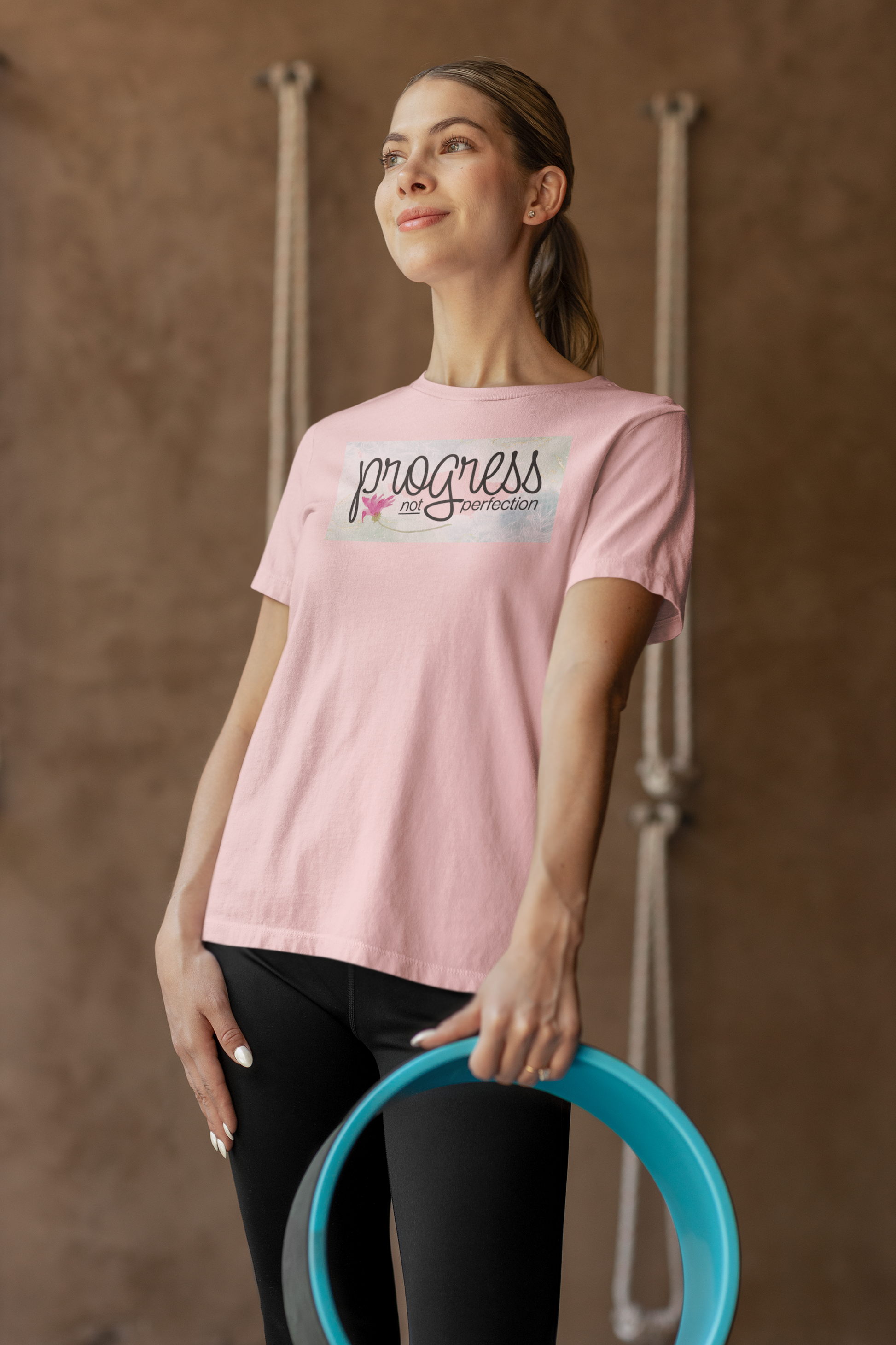 Woman wearing a pink t-shirt with 'progress is the thought' text, holding a blue hoop against a brown background.