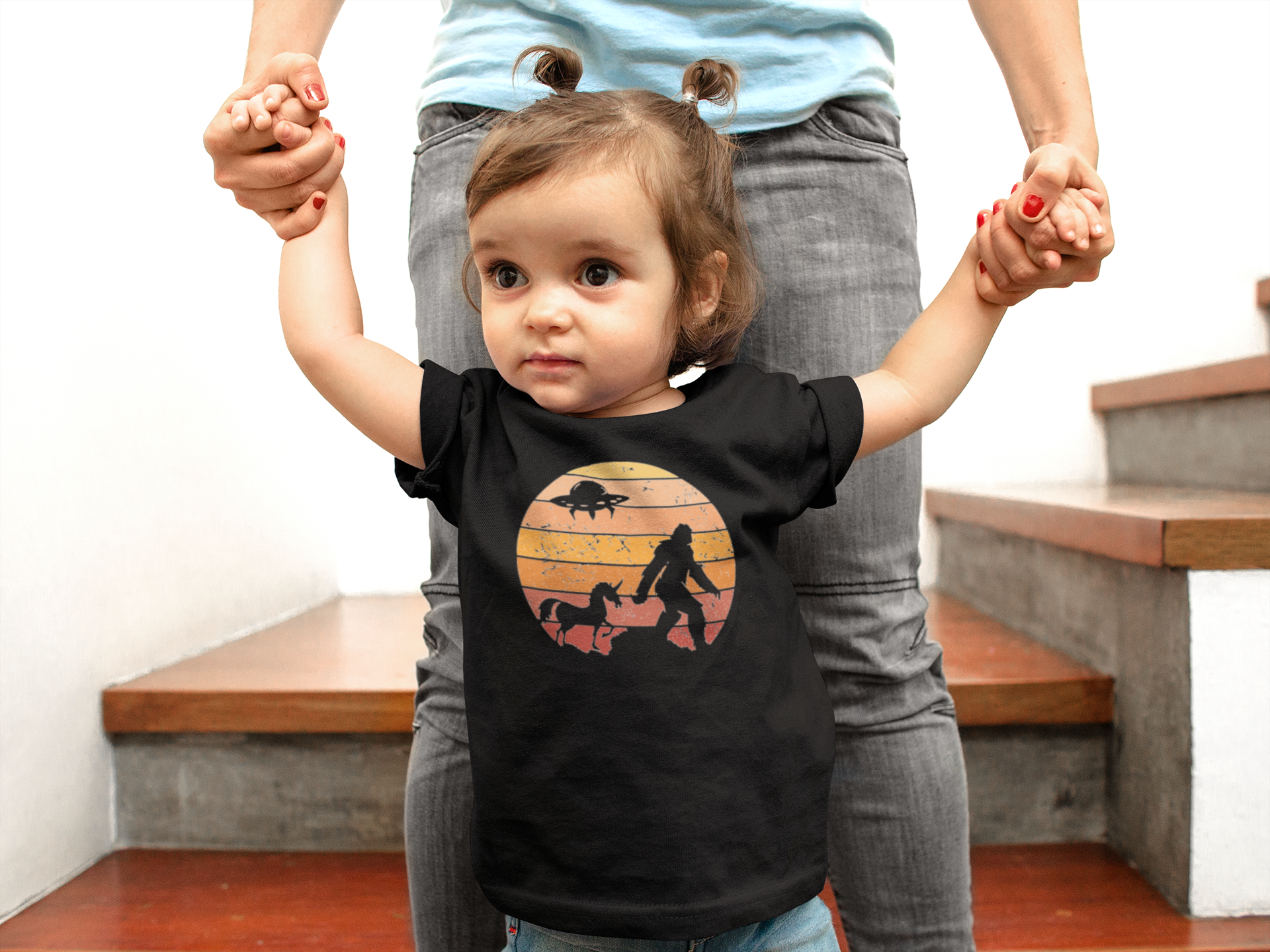 Child wearing a black t-shirt with a bigfoot, ufo and unicorn design, standing with an adult on a staircase.