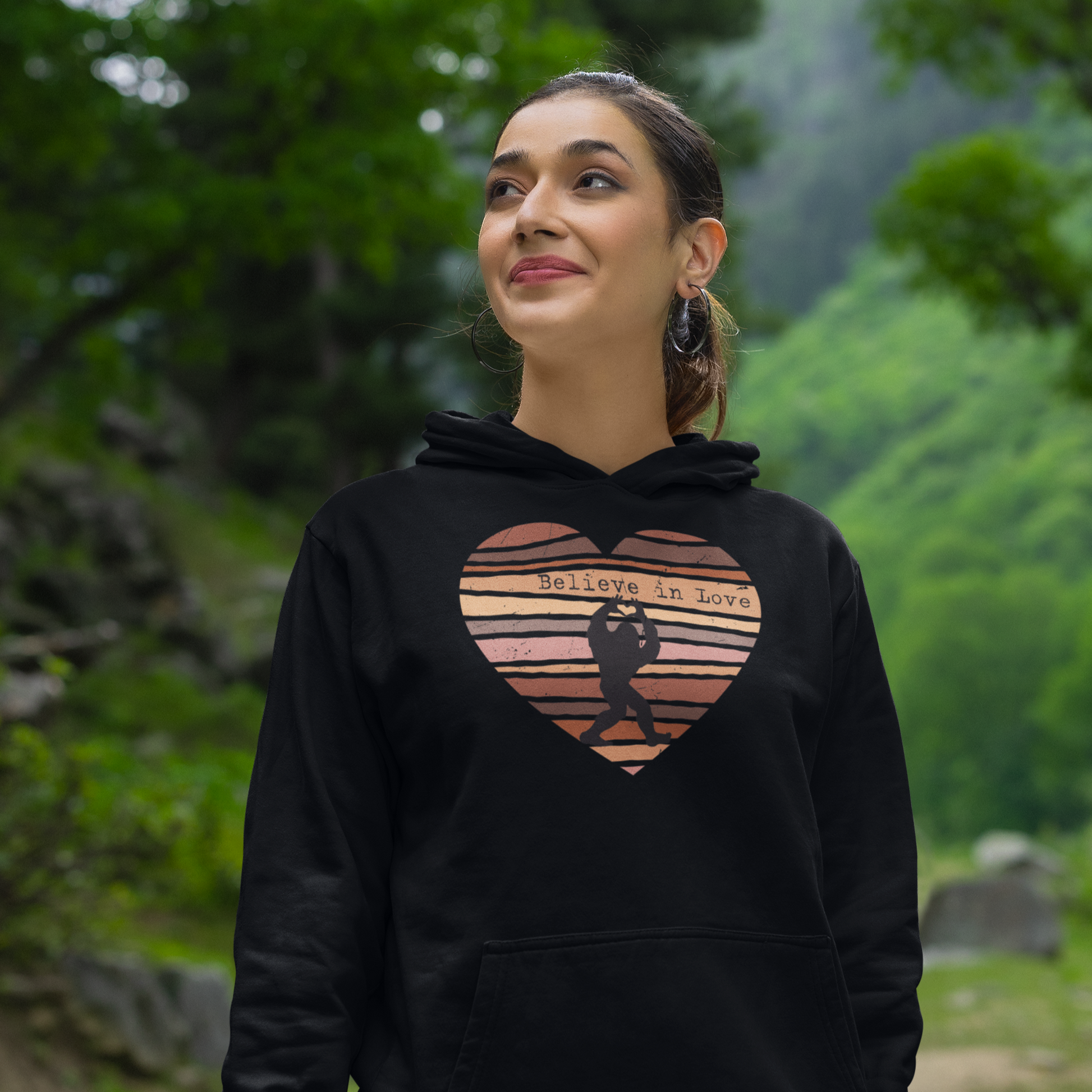 Woman wearing a black hoodie with a bigfoot heart design text Believe in Love in a forest setting