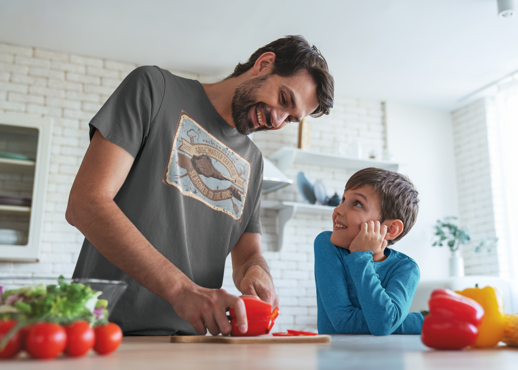 Man and child in a kitchen preparing food together. Man wearing a shirt that says Certified Grill Master. 