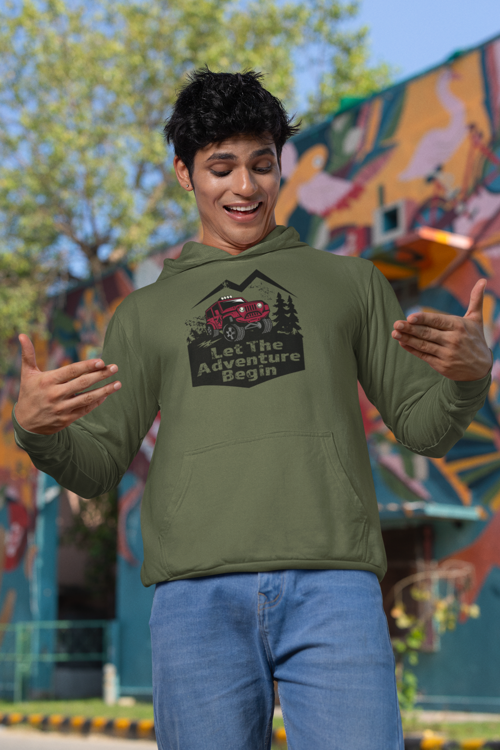 Man wearing a green hoodie with a graphic design that says Let the Adventure begin in front of a colorful mural.
