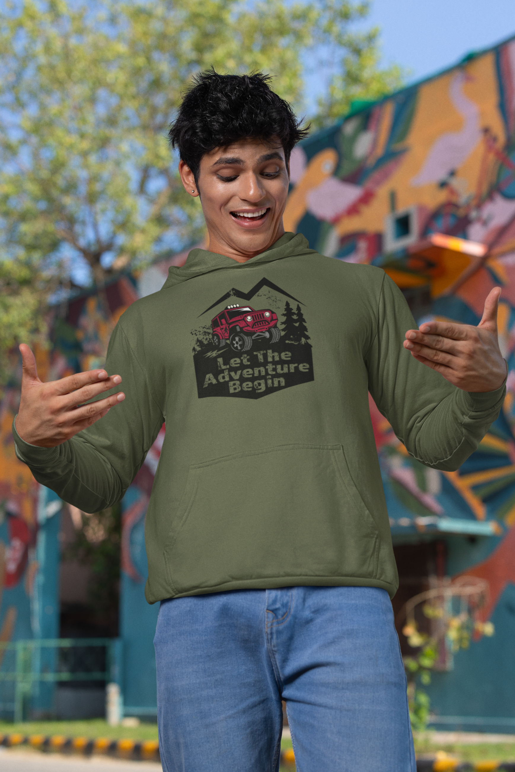 Man wearing a green hoodie with a graphic design that says Let the Adventure begin in front of a colorful mural.