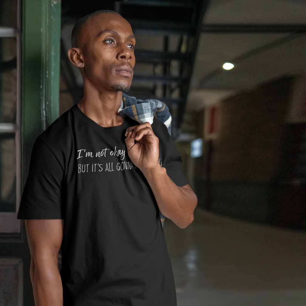 Man wearing a black t-shirt with text, "I'm not okay but it's all gonna be alright" standing in an indoor setting.