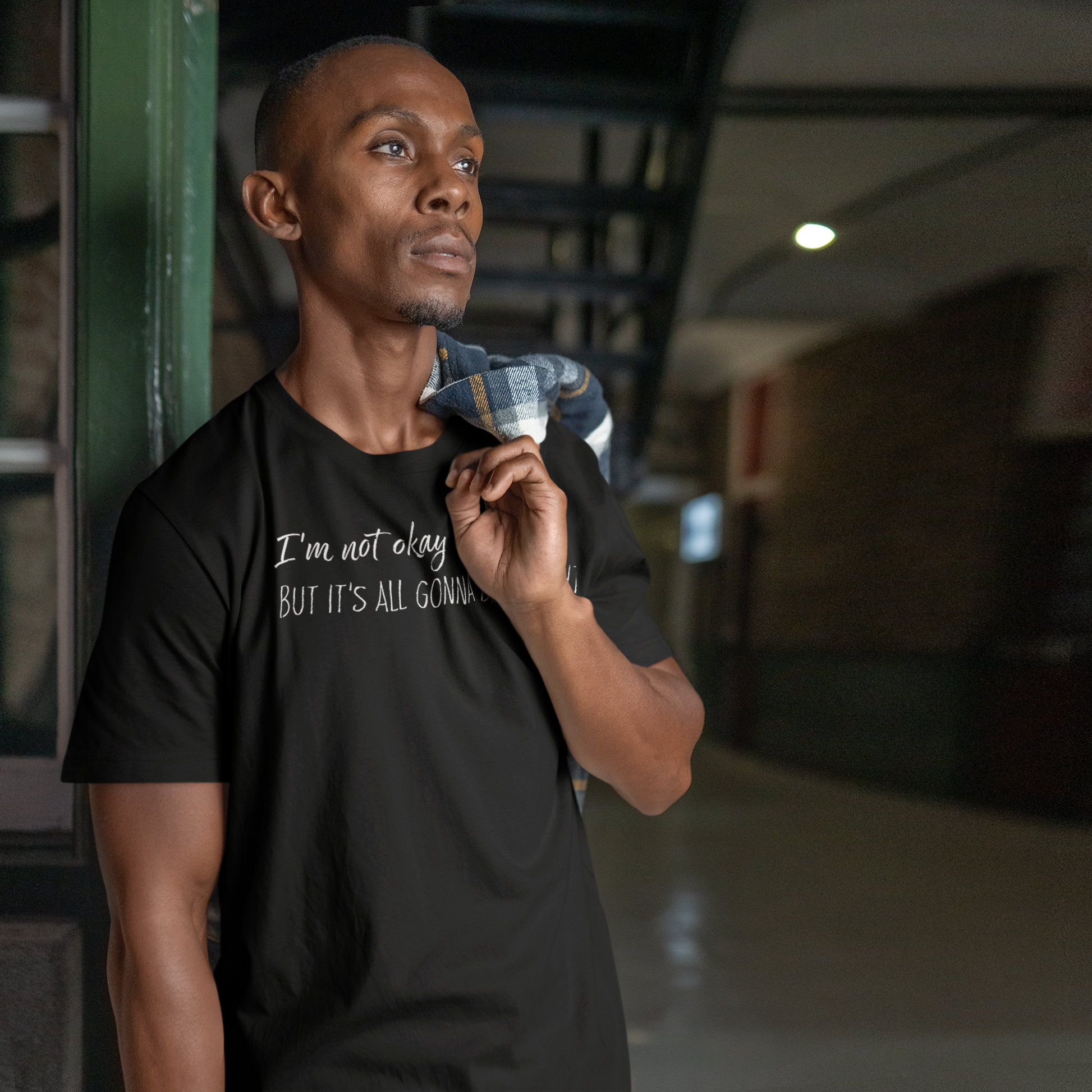 Man wearing a black t-shirt with text, "I'm not okay but it's all gonna be alright" standing in an indoor setting.