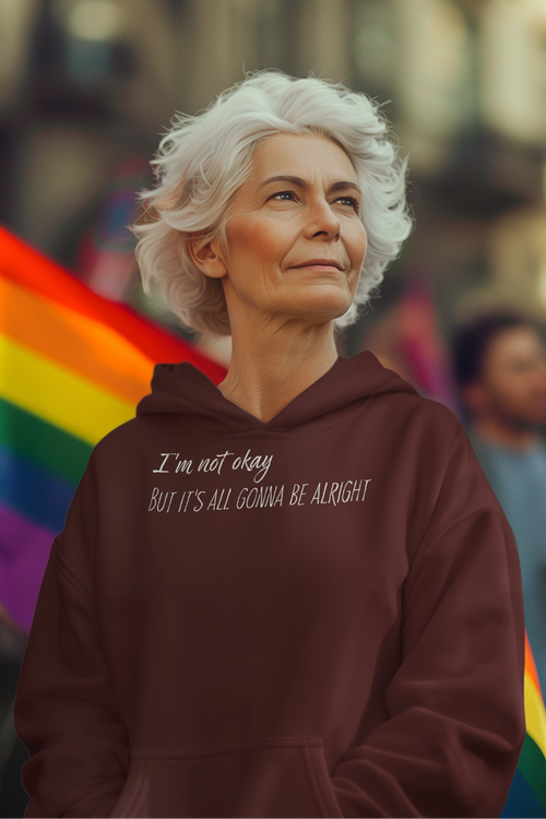 Person wearing a hoodie with text "I'm not okay but it's all gonna be alright", standing in front of rainbow flags.