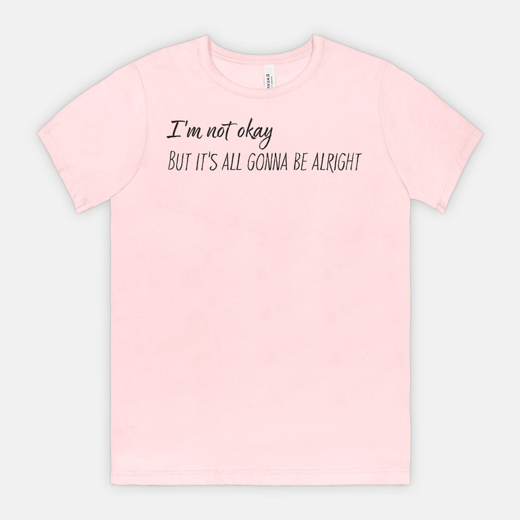 Pink t-shirt with text 'I'm not okay, but it's all gonna be alright' on a light gray background