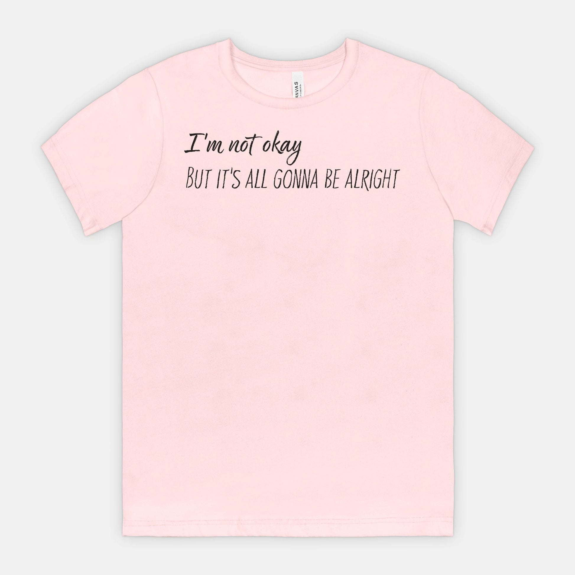 Pink t-shirt with text 'I'm not okay, but it's all gonna be alright' on a light gray background