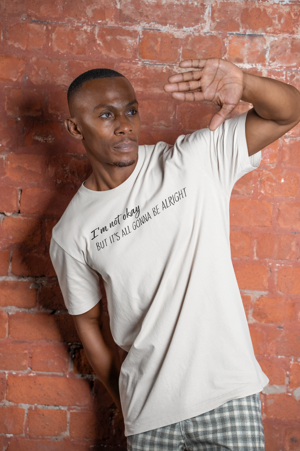 Man wearing a white t-shirt with text "I'm not okay but it's all gonna be alright" against a brick wall
