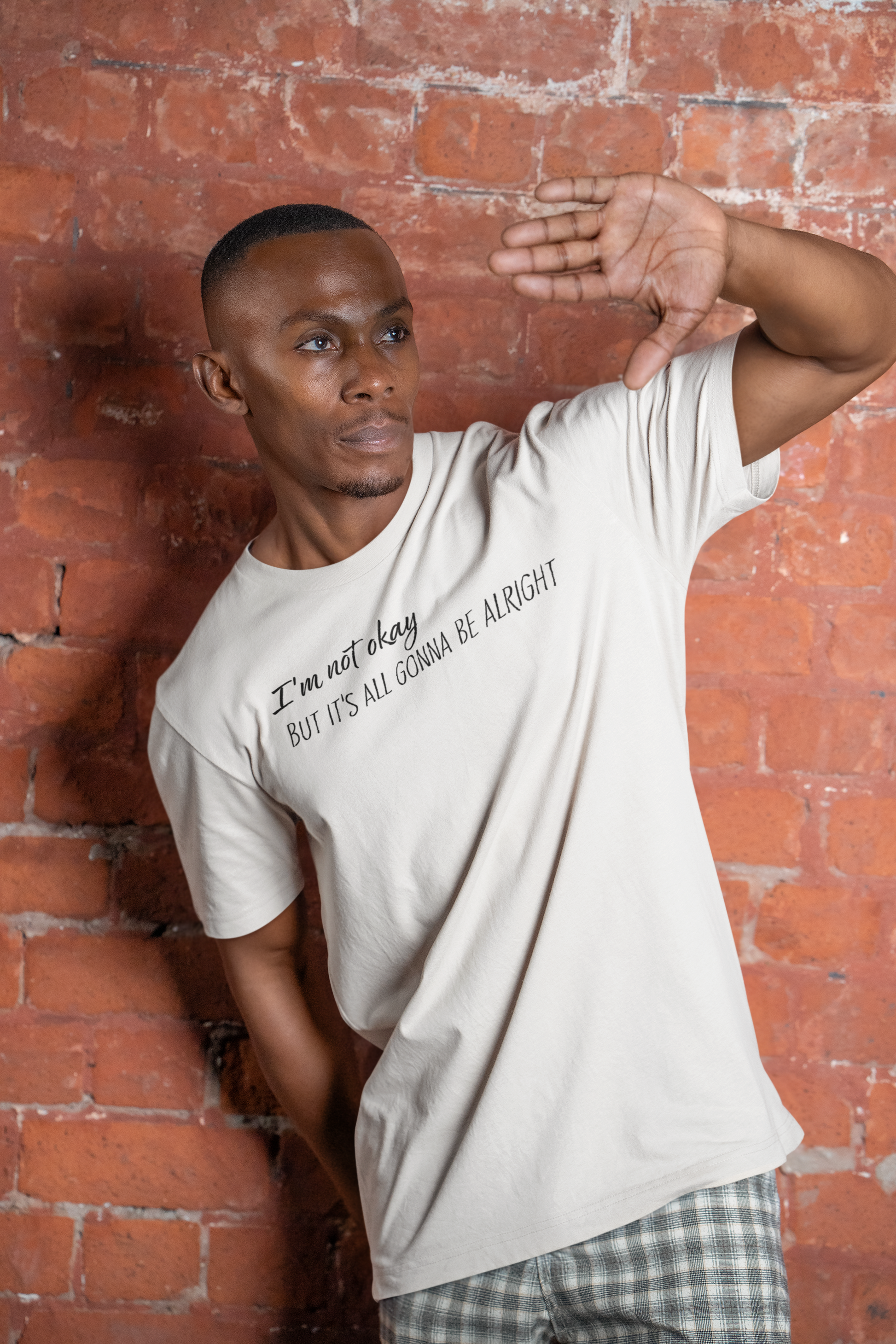 Man wearing a white t-shirt with text "I'm not okay but it's all gonna be alright" against a brick wall