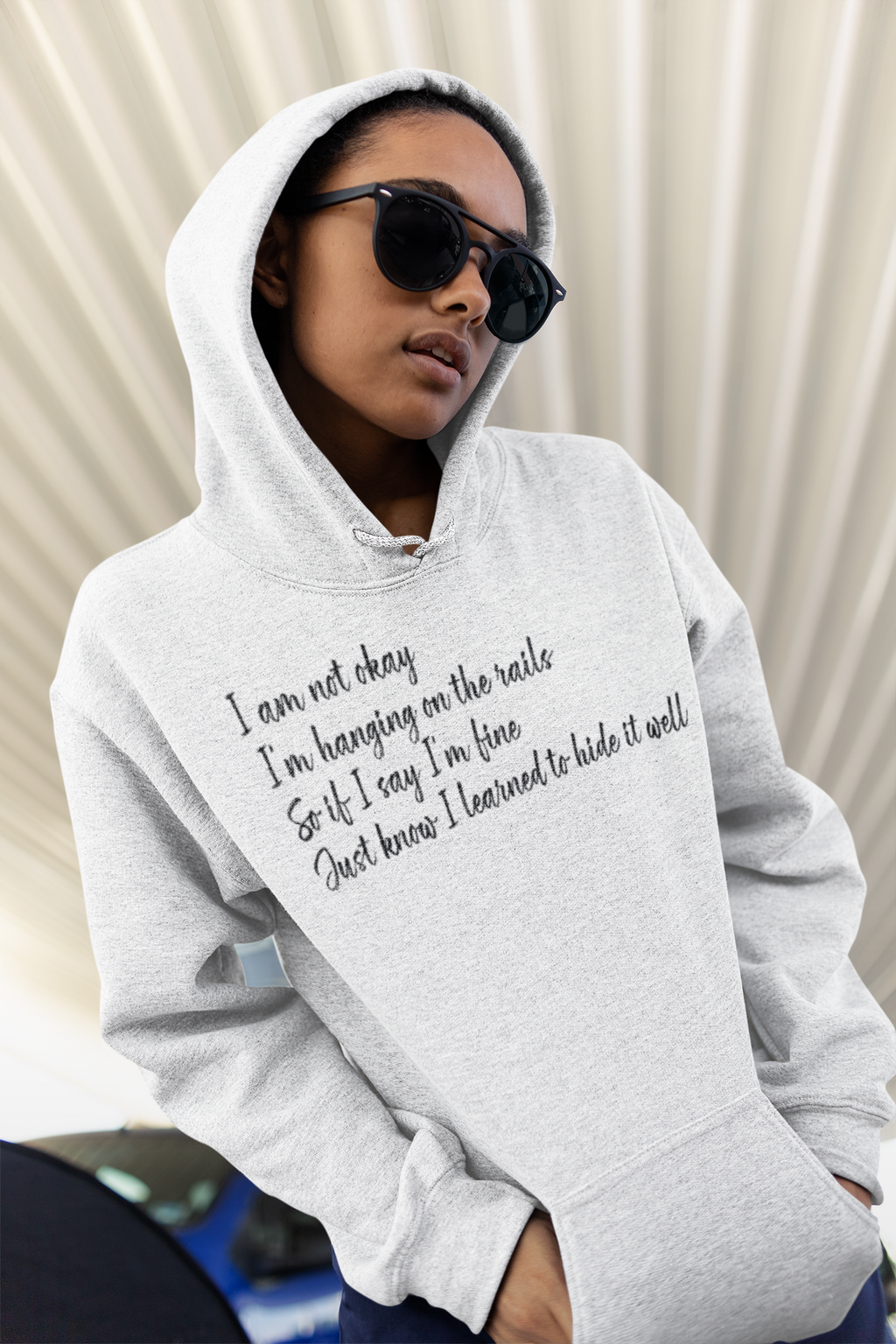 Person wearing a gray hoodie with text, I am not okay, I 'm Hanging on the rails . . . standing against a neutral background