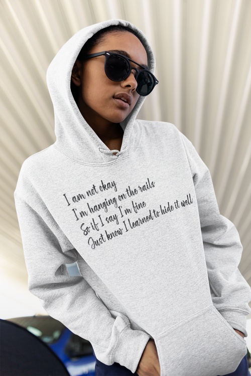 Person wearing a gray hoodie with text, I am not okay, I 'm Hanging on the rails . . . standing against a neutral background