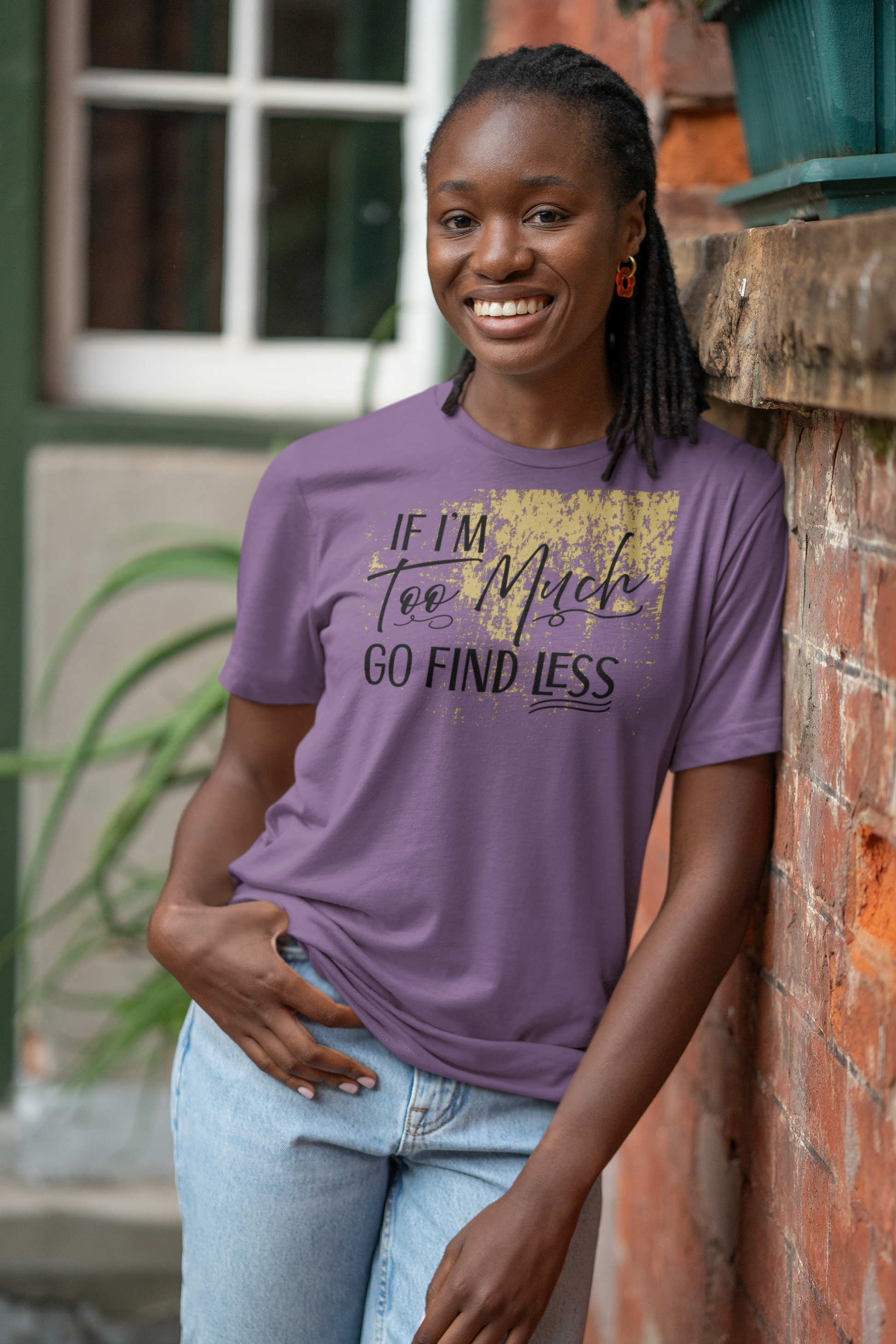 Woman wearing a purple t-shirt with text If I'm Too Much, Go Find Less, standing against a brick wall.
