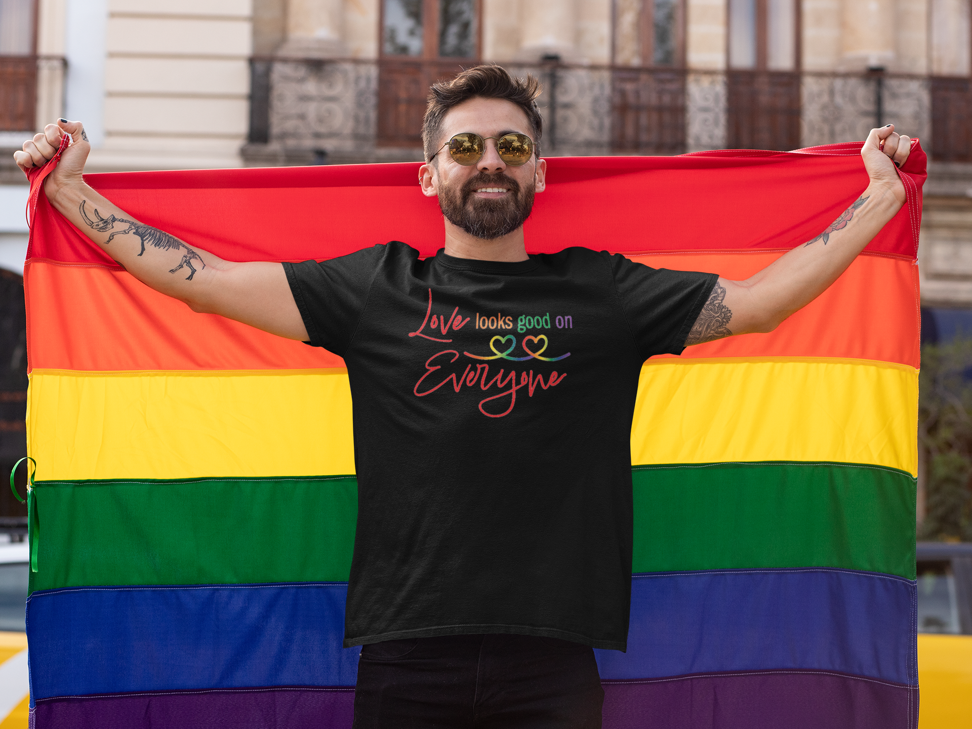 Man holding a rainbow flag with a black t-shirt displaying a message that says Love looks good on Everyone.