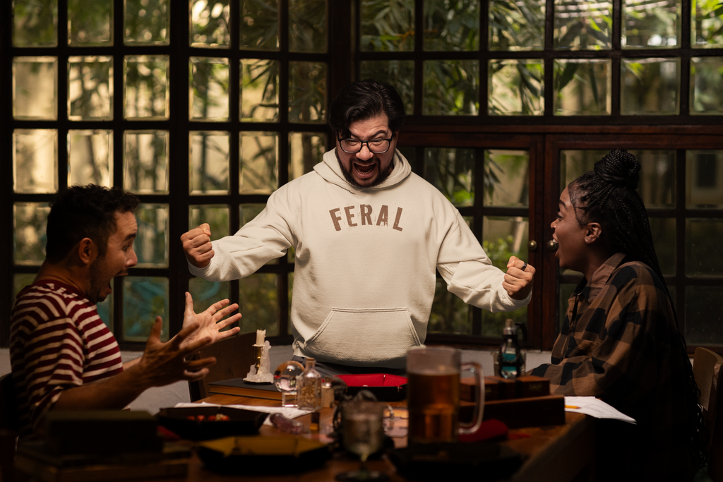 Three people sitting at a table with one person in a 'FERAL' hoodie gesturing animatedly. Gildan 18500 sand hoodie featuring the word “FERAL” in bold distressed brown lettering, designed by Red Rock Chaos for a minimalist wild-at-heart style.