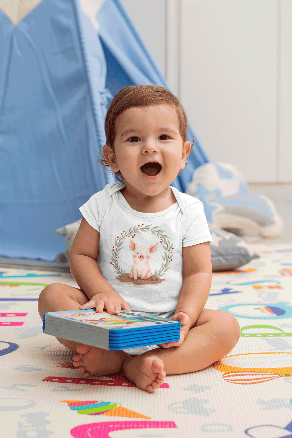 Child sitting on a colorful mat holding a book with a blue tent in the background wearing Little Chaos Crew piglet sitting in a mud puddle with eyes closed inside green leafy wreath, minimalist baby onesie white design.