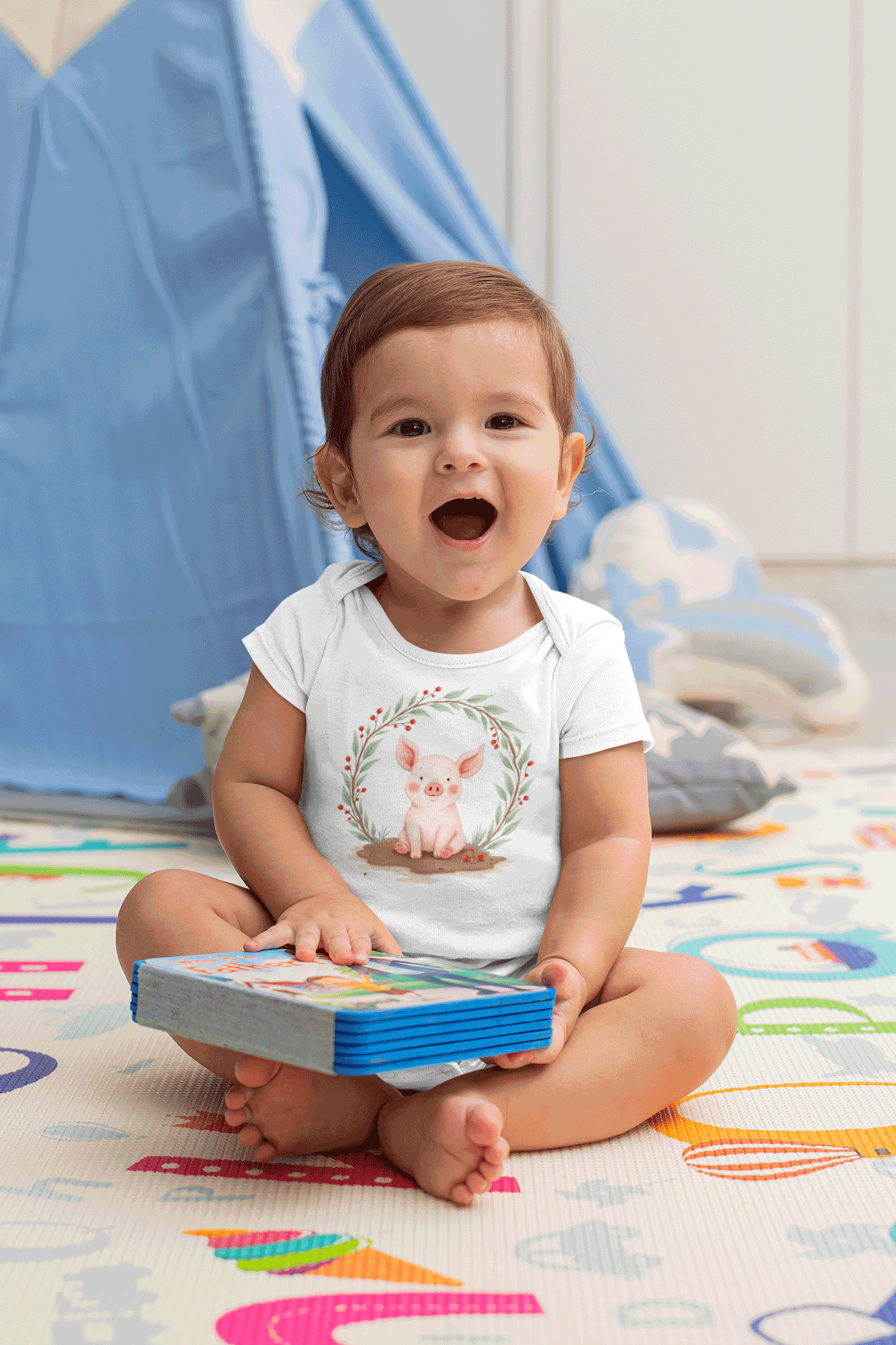 Child sitting on a colorful mat holding a book with a blue tent in the background wearing Little Chaos Crew piglet sitting in a mud puddle with eyes closed inside green leafy wreath, minimalist baby onesie white design.