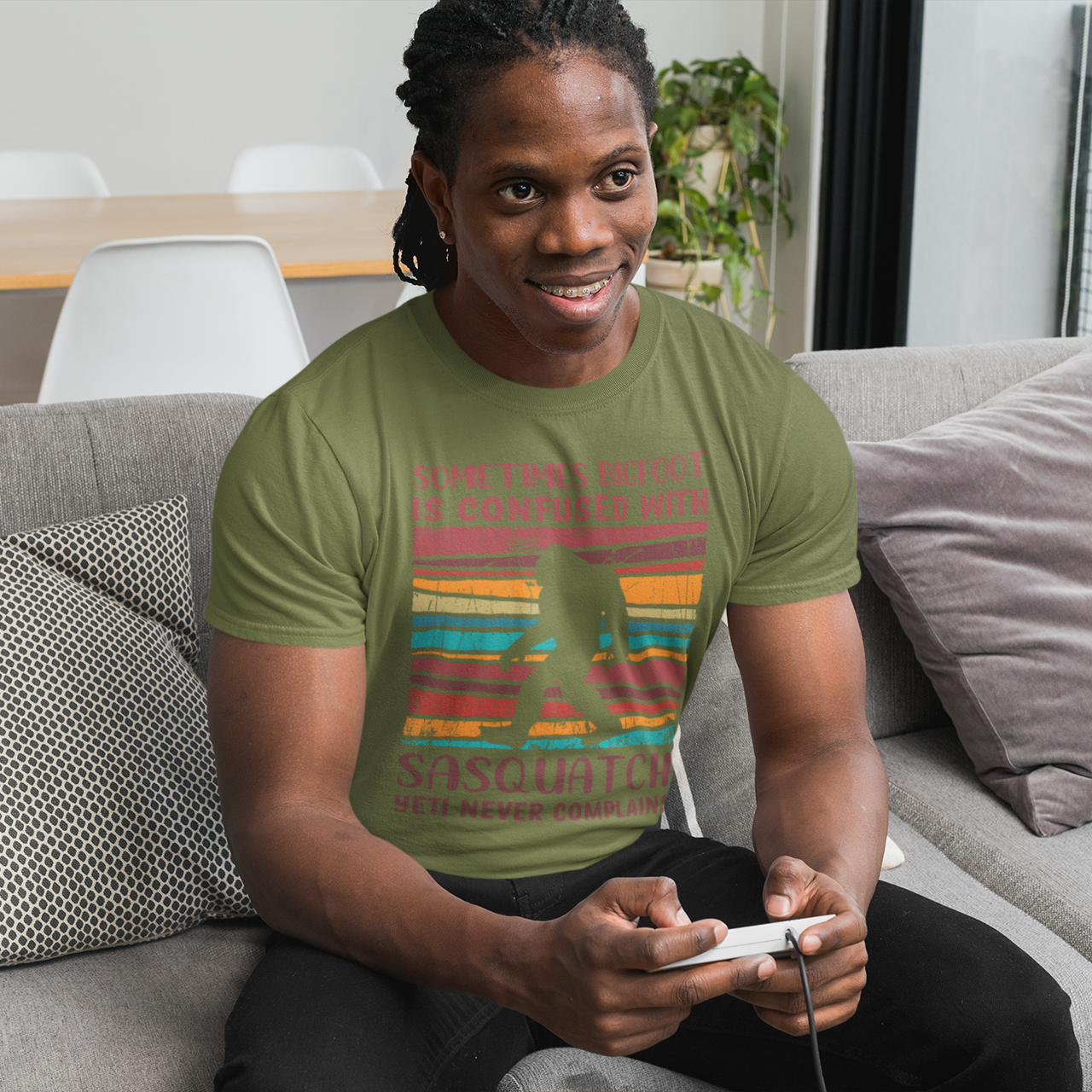 Person sitting on a couch wearing a green t-shirt with colorful text Sometimes Bigfoot is confused with Sansquatch Yeti never Complains, holding a remote control.